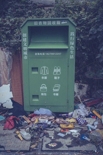 A green, metal clothing donation bin positioned on a concrete pavement, set against a background of dense foliage and a wooden fence. The bin has Chinese characters on the top and sides, indicating it